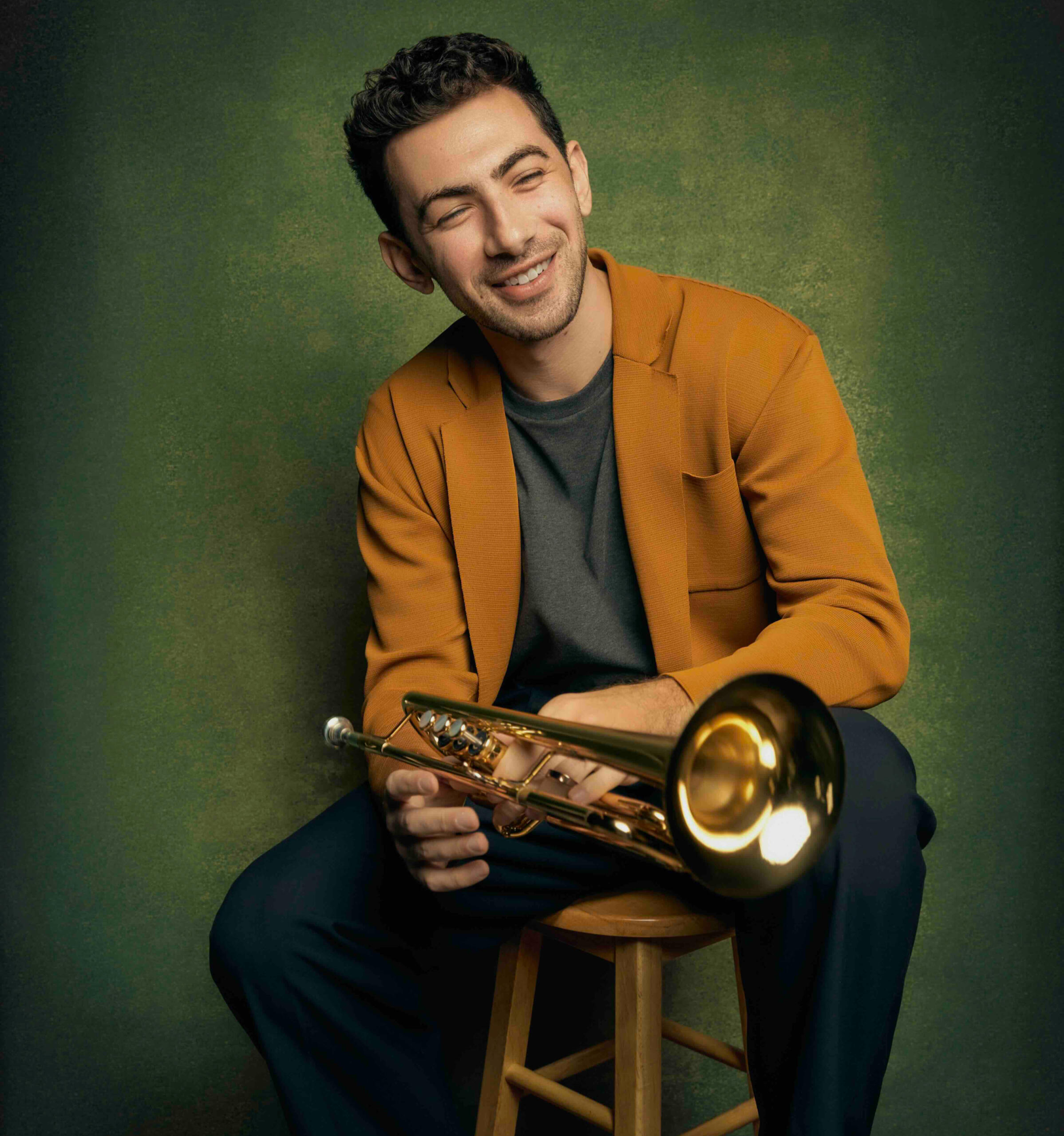 Earth Tones Trumpet Stool Musician John Dimase sitting wearing autumnal colors and holding his trumpet with a gentle smile. Sitting on a stool. Green backdrop.