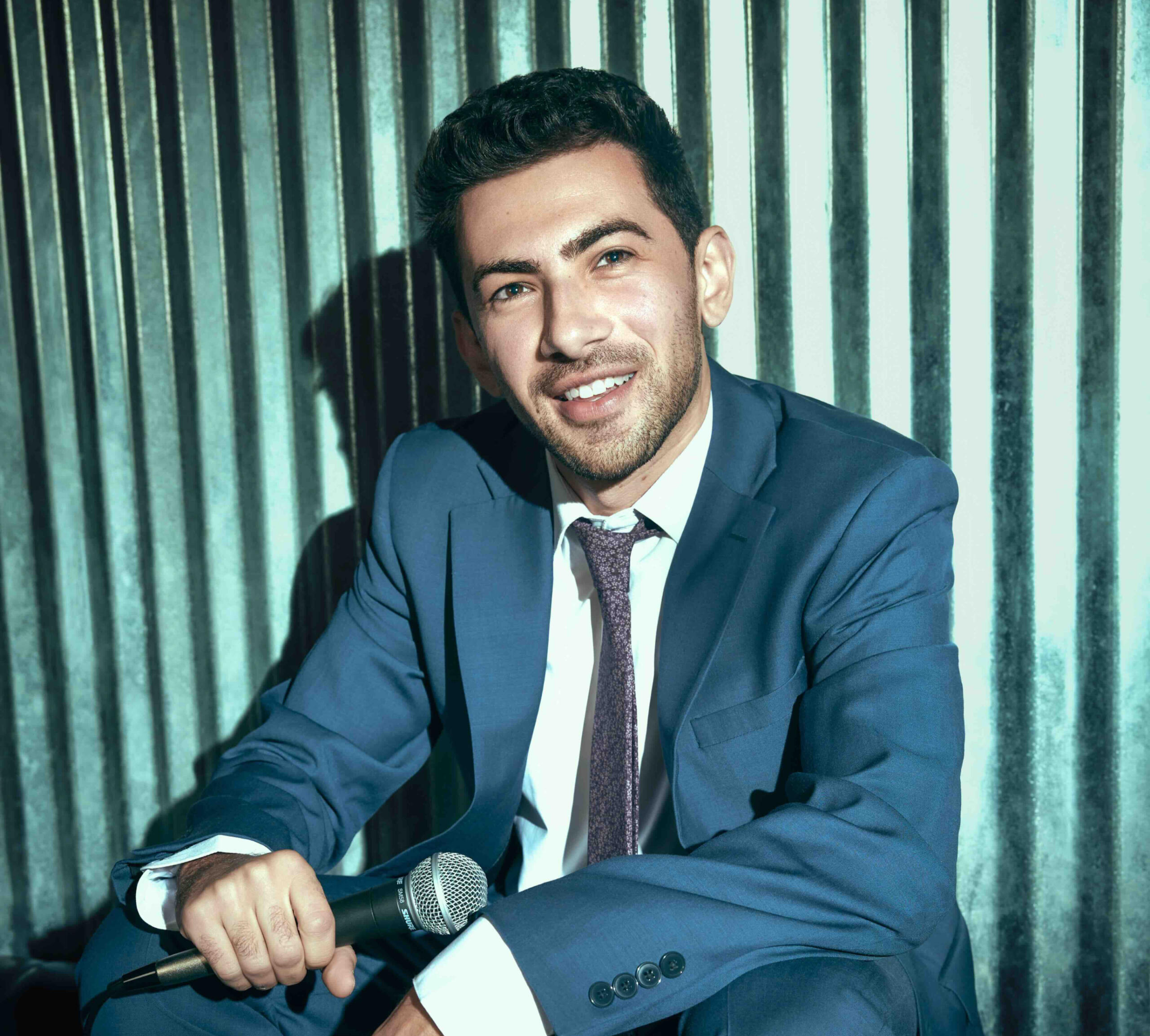 Suit Couch Sitting Musician John Dimase smiling while wearing a blue suit, white shirt, purple tie. Metal backdrop and cool color tones make the image "pop".